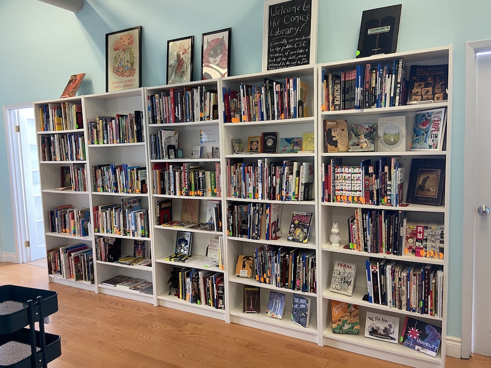 A sneak peek at our new cozy space on the 4th floor of @csitoronto Annex! Lots of colourful comics on tall white shelves surrounded by light blue walls, with several larger comics displayed on top of the shelves along with framed artwork and a chalkboard sign that reads “Welcome to the comics library!”